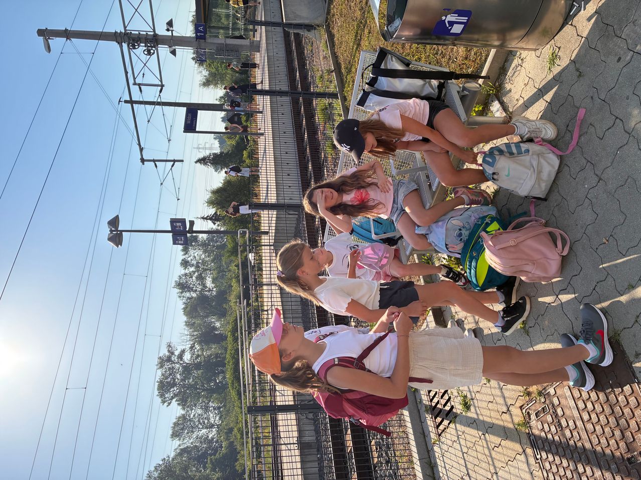 Eine Gruppe von Mädchen mit Rucksäcken wartet auf einem Bahnsteig. Sie sitzen auf dem Boden und lehnen sich gegen einen Zaun. Bäume und Stromleitungen sind im Hintergrund.