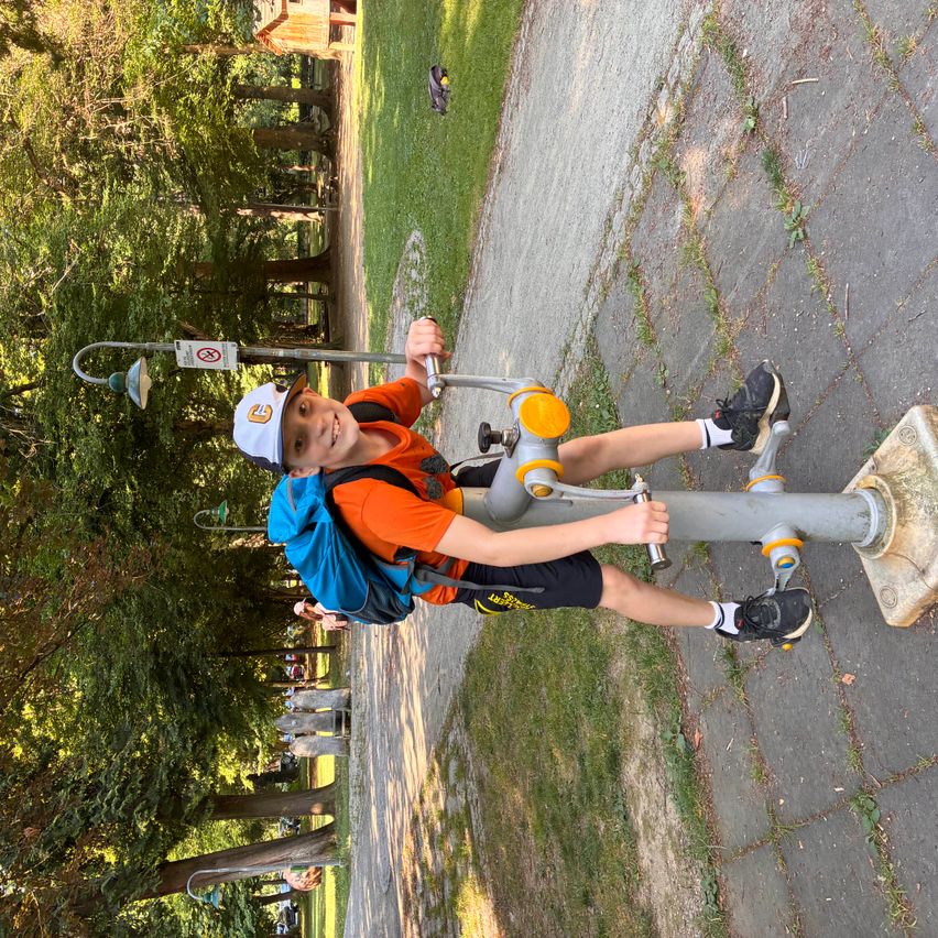 Ein junger Junge benutzt ein Outdoor-Trainingsgerät in einem Park. Er trägt ein orangefarbenes T-Shirt, schwarze Shorts und Turnschuhe. Er hat einen blauen Rucksack.
