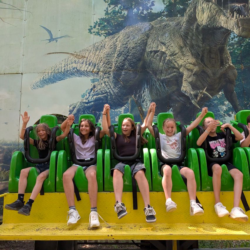Five children sit in a roller coaster ride, wearing sneakers. The ride has green seats with yellow base, and the background is a dinosaur painting.