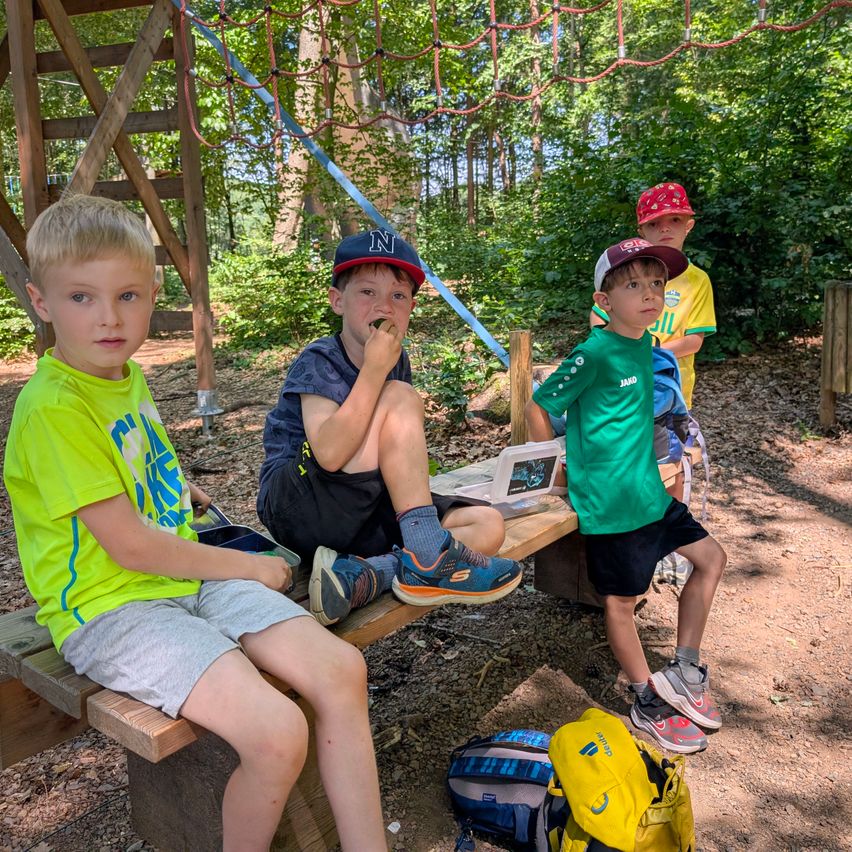 Four boys in hats sit on a wooden bench in a forest. One boy eats a snack. A backpack and a tablet are in front of them.