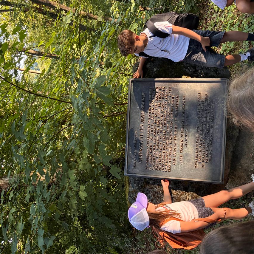 Kinder und eine Frau betrachten eine Gedenktafel in der Nähe eines Waldweges. Die Tafel erinnert an Dr. Josef Steiner, mit eingravierten Daten und Namen.