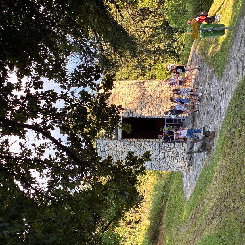 Eine Gruppe von Menschen steht vor einem steinernen Gebäude mit einem grünen Mülleimer an der Seite. Sie tragen Turnschuhe.