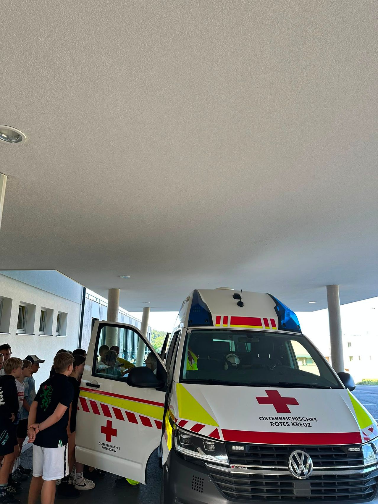 A white ambulance with red crosses on its side and roof is parked at a building. A group of children is gathered near the ambulance, possibly waiting for it to leave.
