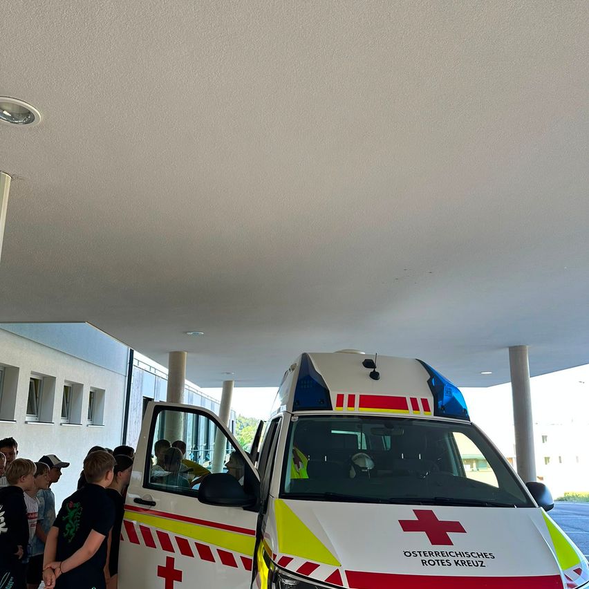 Ein weißer Krankenwagen mit roten Kreuzen auf der Seite und dem Dach ist an einem Gebäude geparkt. Eine Gruppe von Kindern steht in der Nähe des Krankenwagens, möglicherweise wartend, dass er wegfährt.
