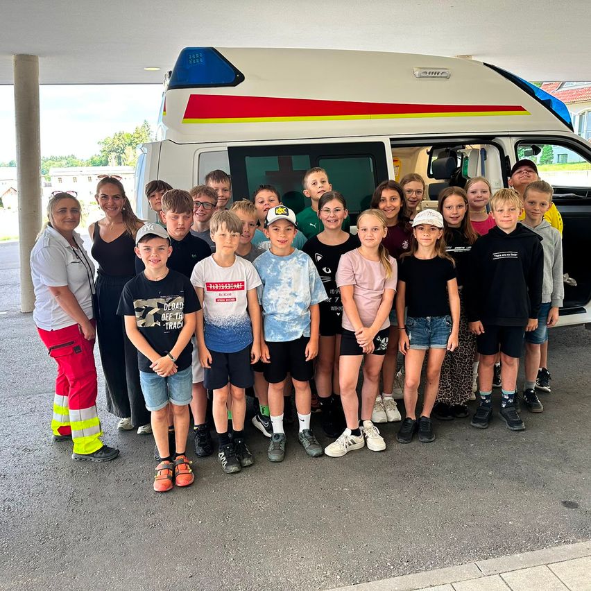 A group of children and adults pose in front of an ambulance with a woman in reflective clothing.