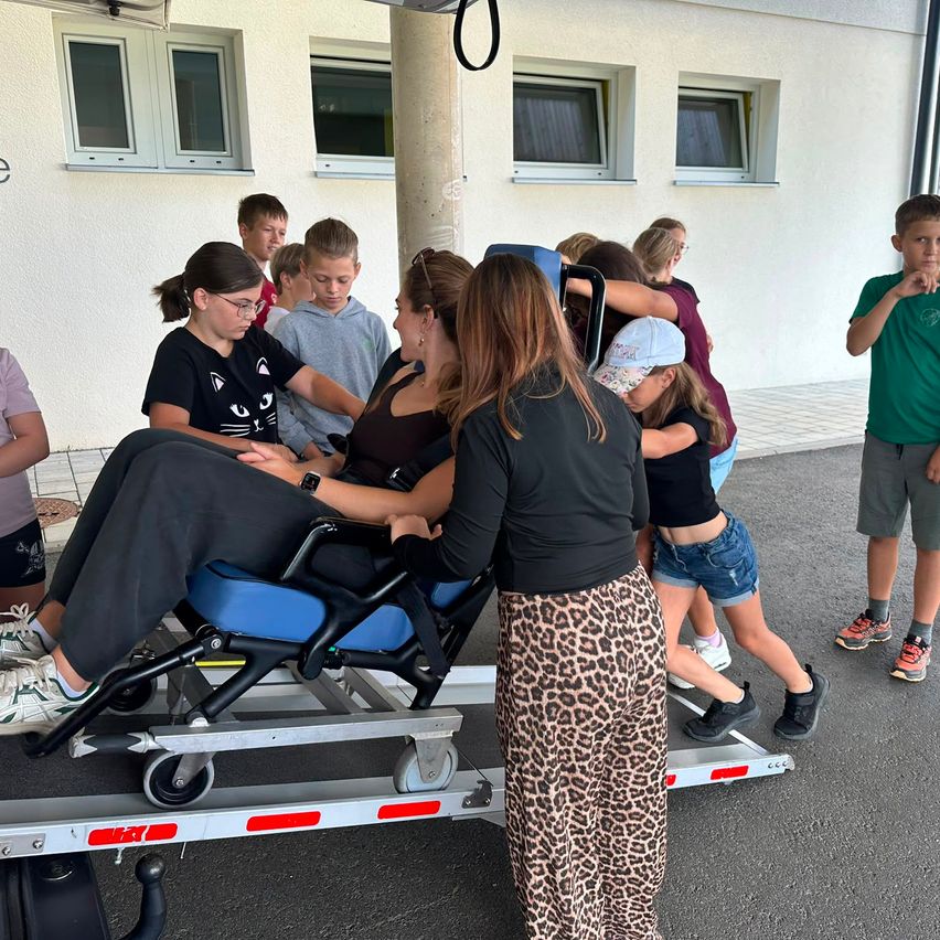 A group of children and two women stand around a woman sitting on a wheelchair. The wheelchair is on a trailer, with a woman standing next to it. They are outside in front of a building.