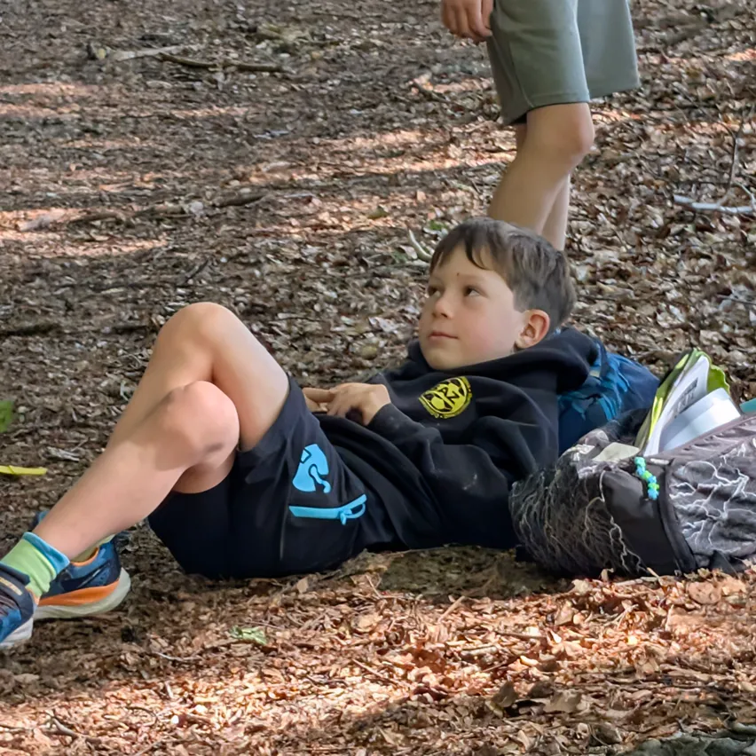 Ein junger Junge liegt auf dem Boden in einem Wald, trägt eine schwarze Kapuzenjacke mit einem gelben Logo und blaue Shorts. Er schaut zur Seite, möglicherweise für ein Foto posierend.