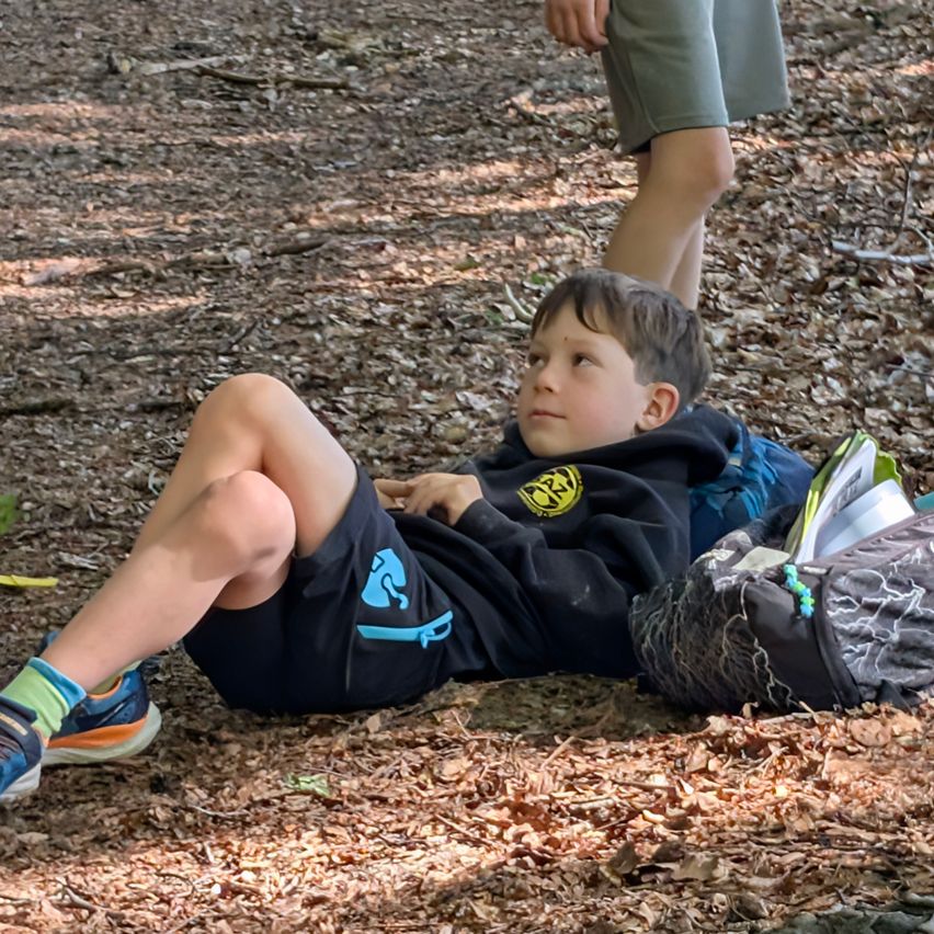 Ein junger Junge liegt auf dem Boden in einem Wald, trägt eine schwarze Kapuzenjacke mit einem gelben Logo und blaue Shorts. Er schaut zur Seite, möglicherweise für ein Foto posierend.