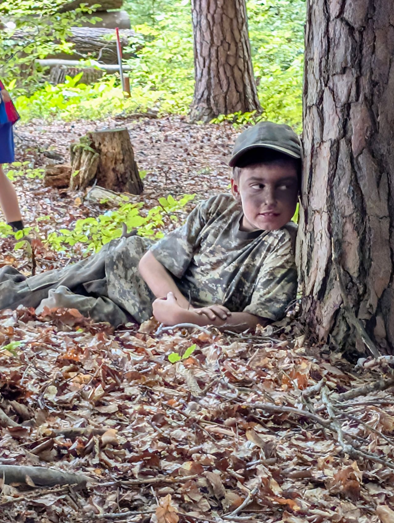 A young boy in camouflage clothing lies on the ground in a forest, leaning against a tree.