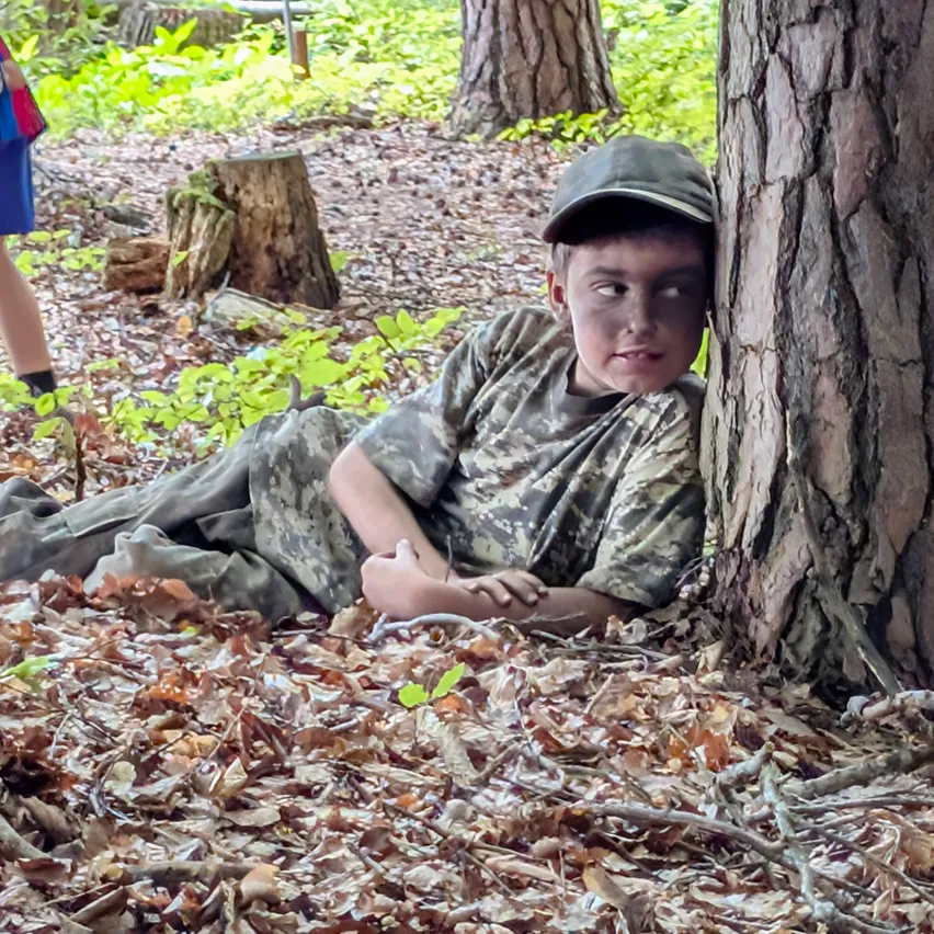 Ein Junge in Tarnkleidung liegt auf dem Boden in einem Wald und lehnt sich gegen einen Baum.