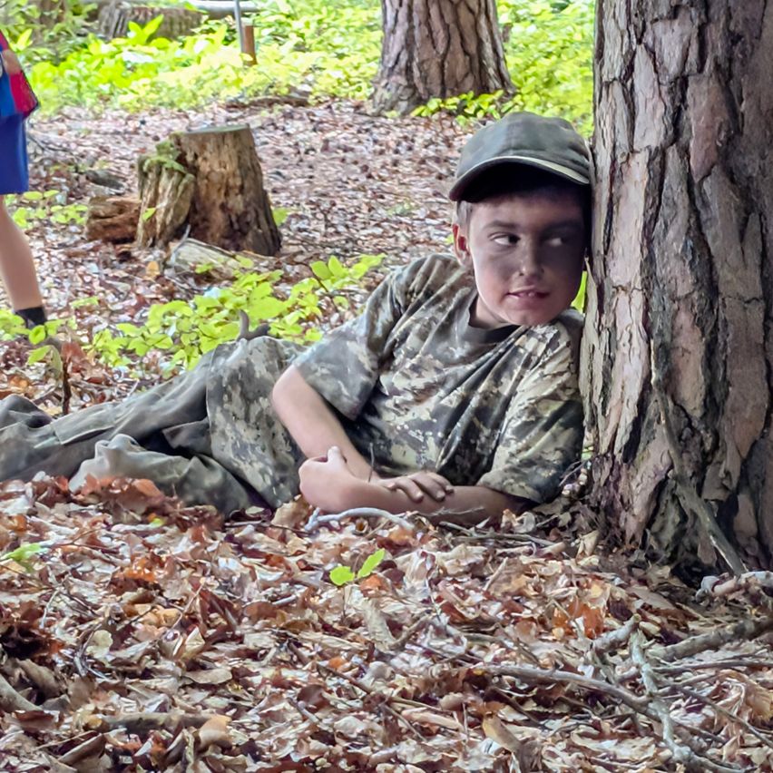 Ein Junge in Tarnkleidung liegt auf dem Boden in einem Wald und lehnt sich gegen einen Baum.