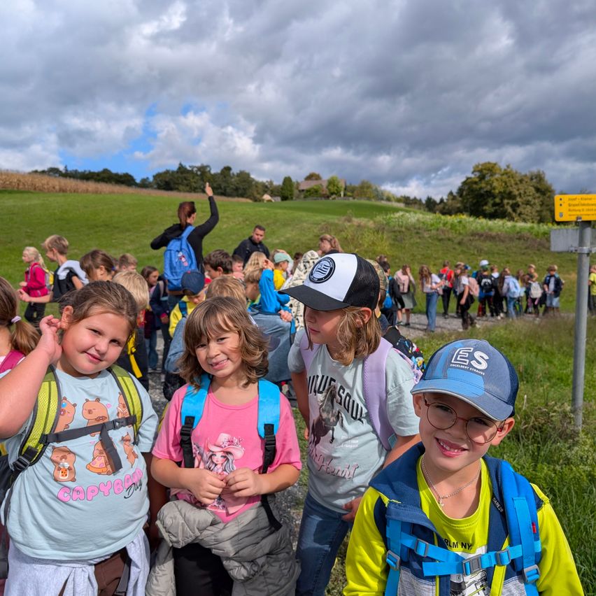 Eine Gruppe von Kindern auf einer Wiese, einige mit Rucksäcken, lächeln und posieren für ein Foto. Ein Schild ist im Hintergrund zu sehen.