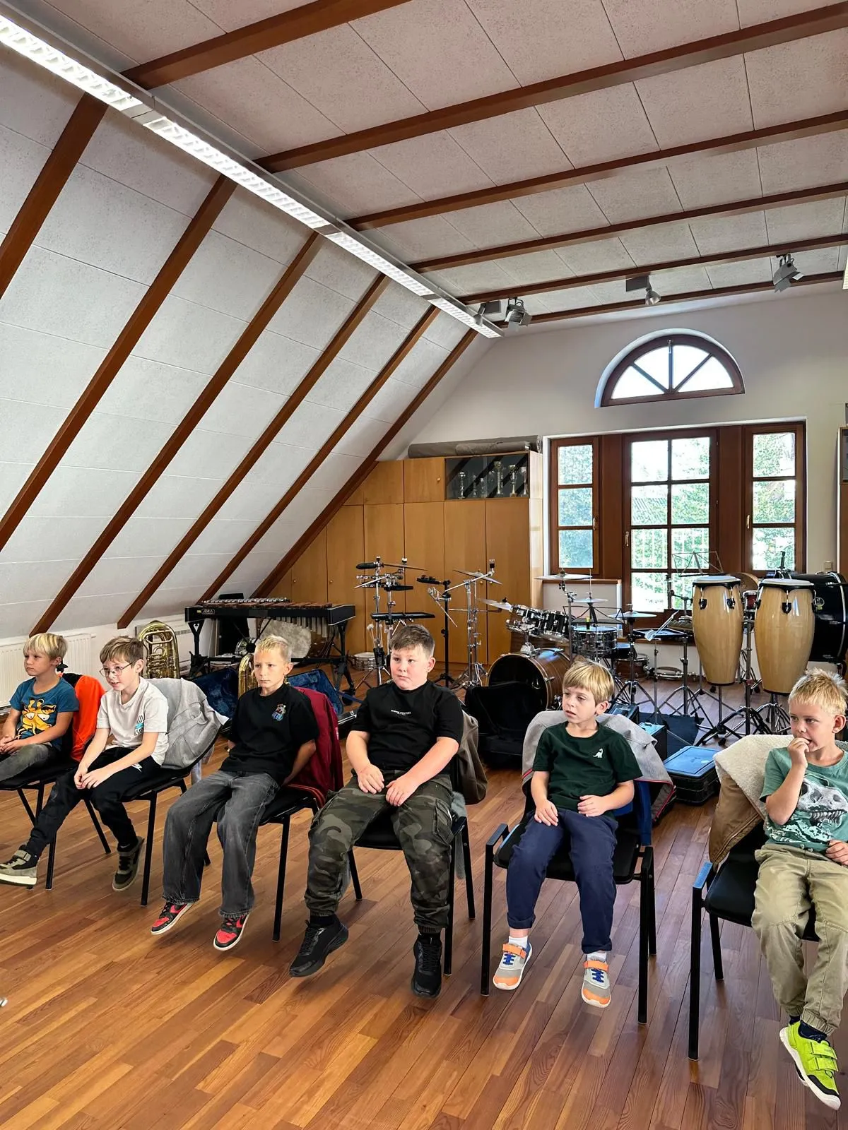 Eine Gruppe junger Jungen sitzt in Stühlen in einem Musikraum mit verschiedenen Musikinstrumenten um sie herum.