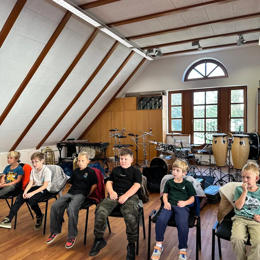 Eine Gruppe junger Jungen sitzt in Stühlen in einem Musikraum mit verschiedenen Musikinstrumenten um sie herum.