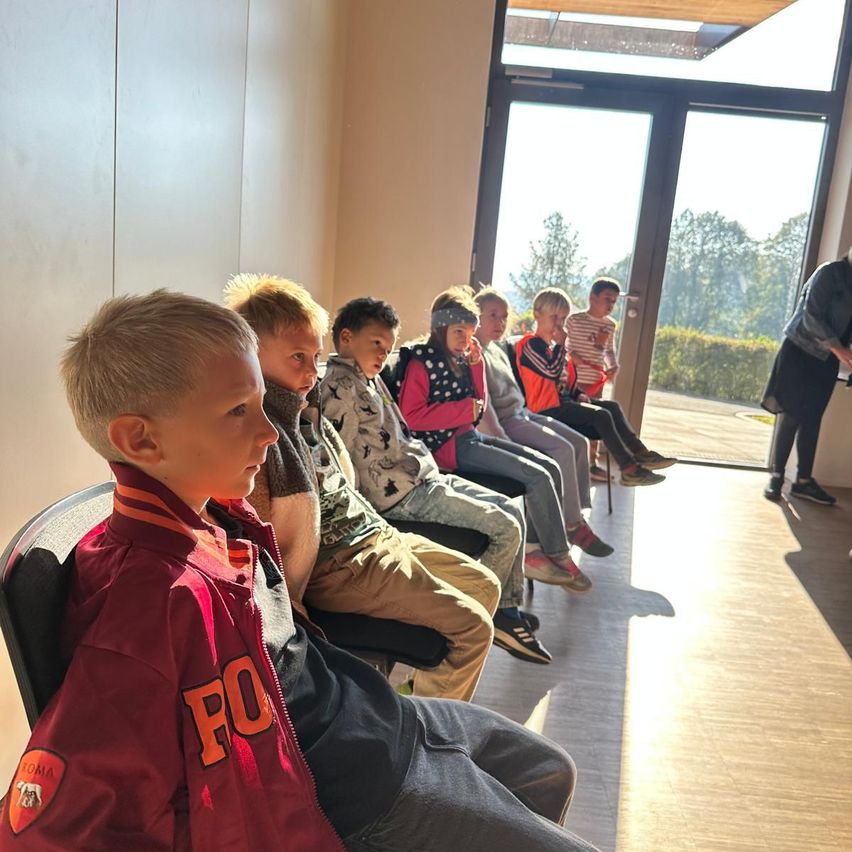 Eine Gruppe von Kindern sitzt in Stühlen in einem Raum mit Glastüren und schaut auf Bäume und eine Person, die in der Nähe steht.