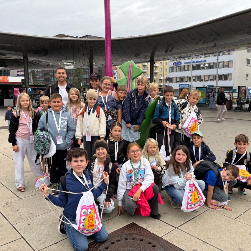 Eine Gruppe von Kindern und Erwachsenen posiert für ein Foto vor einer großen grünen Skulptur. Sie tragen alle Ausweise und einige haben Taschen. Der Ort ist im Freien mit einem modernen Gebäude im Hintergrund.
