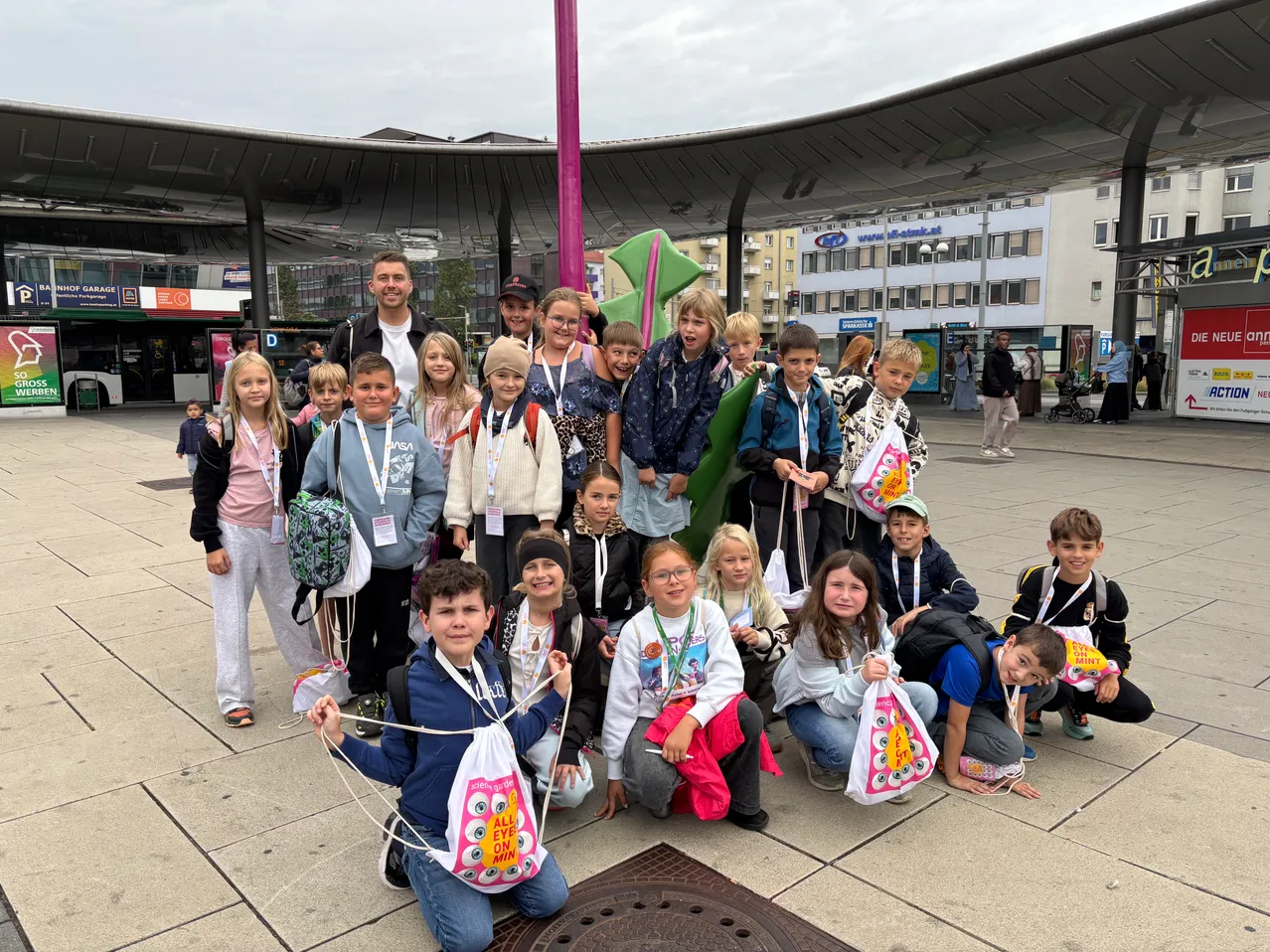 Eine Gruppe von Kindern und Erwachsenen posiert für ein Foto vor einer großen grünen Skulptur. Sie tragen alle Ausweise und einige haben Taschen. Der Ort ist im Freien mit einem modernen Gebäude im Hintergrund.