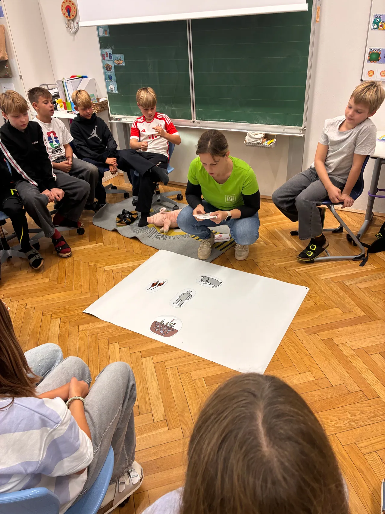 Eine Gruppe von Kindern sitzt im Kreis um eine Frau, die auf dem Boden kniet und ein Spielbrett mit Bildern von Menschen und Tieren hält.