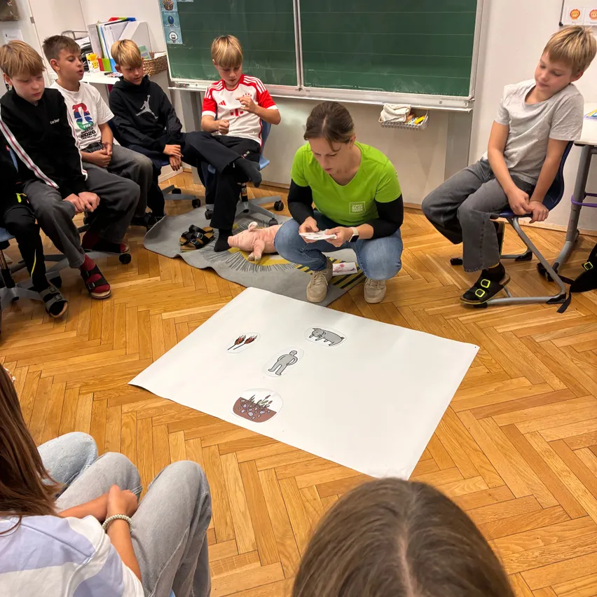 Eine Gruppe von Kindern sitzt im Kreis um eine Frau, die auf dem Boden kniet und ein Spielbrett mit Bildern von Menschen und Tieren hält.