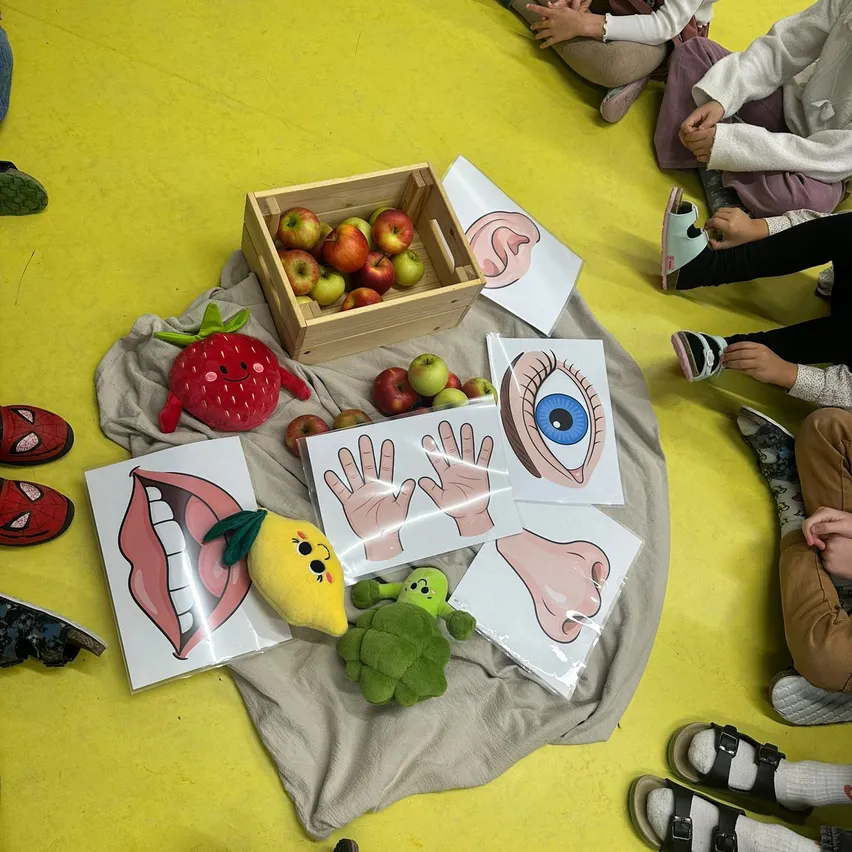 Eine Gruppe von Kindern sitzt auf einem gelben Boden mit verschiedenen Lernmaterialien um sie herum. Es gibt eine Box mit Äpfeln, eine Erdbeer-Plüschfigur und mehrere Zeichnungen von Körperteilen.