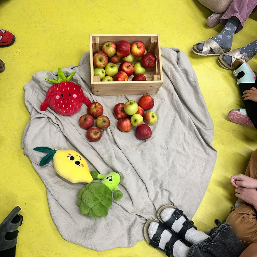 Eine Gruppe von Kindern sitzt auf einem gelben Boden mit verschiedenen Spielzeugen und Früchten. Es gibt eine Holzkiste voller Äpfel, ein Erdbeer-Plüschtier und ein Zitronen-Plüschtier.