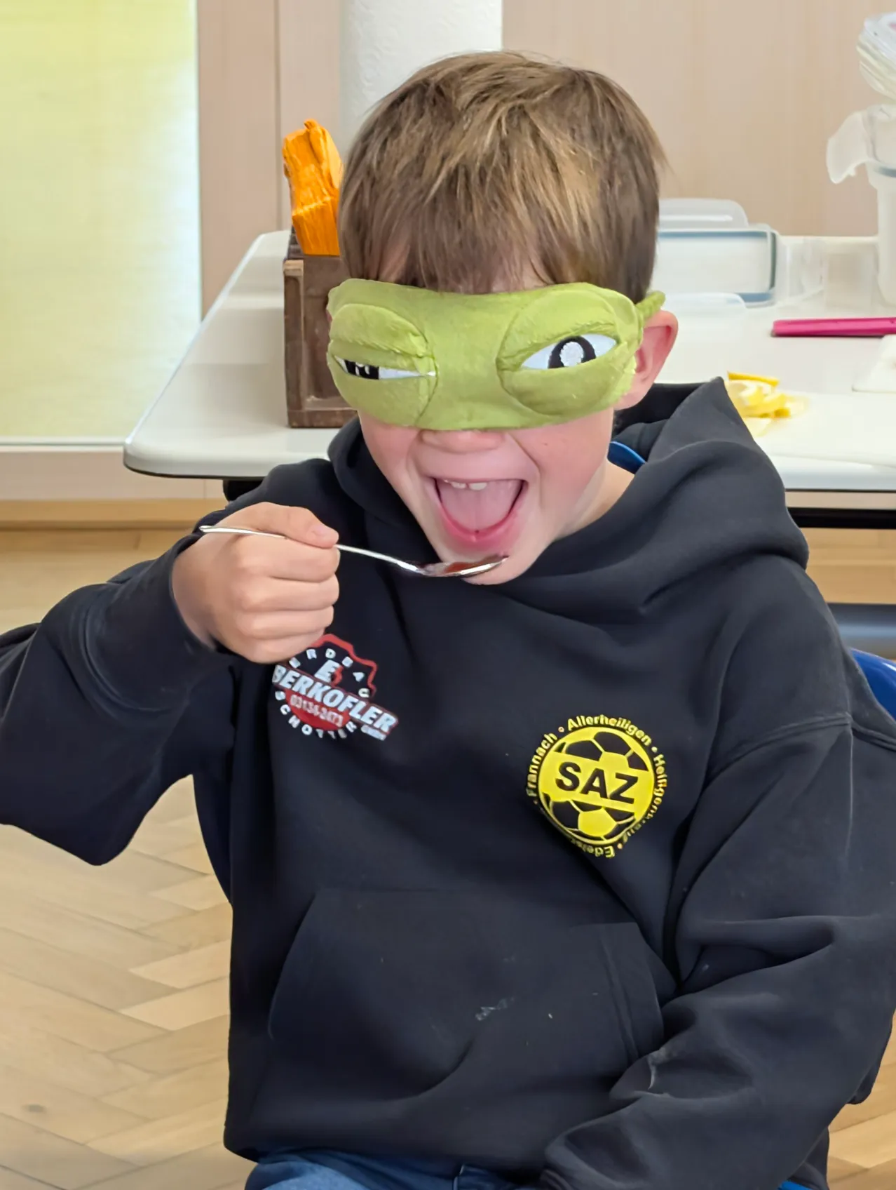 Ein junger Junge mit einem grünen Blindfold isst etwas mit einem Löffel.