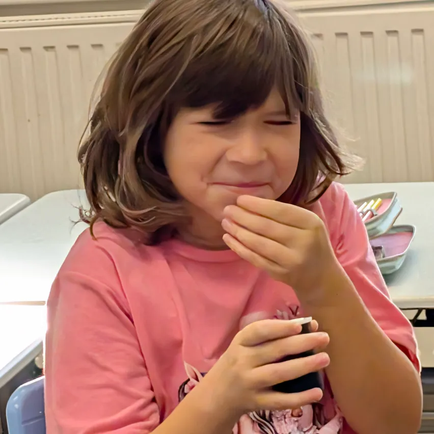Ein junges Mädchen in einem rosa T-Shirt sitzt in einem Klassenzimmer, hält ein Handy an den Mund und lächelt.