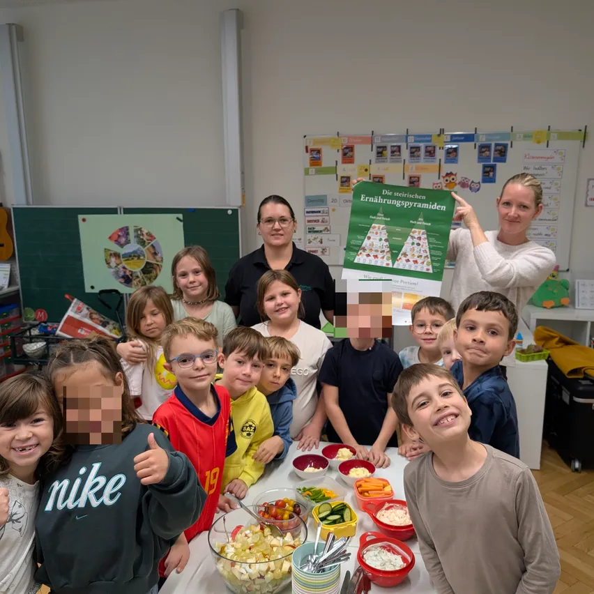 Eine Gruppe von Kindern und zwei Frauen versammeln sich in einem Klassenzimmer. Die Kinder lächeln und posieren für ein Foto. Eine der Frauen hält ein grünes Plakat mit einer Nahrungspyramide.