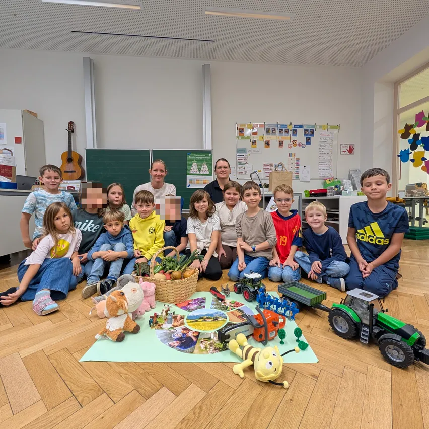 Eine Gruppe von Kindern und zwei Frauen sitzen auf dem Boden in einem Klassenzimmer. Es gibt Spielzeug, einen Korb mit Früchten und ein Poster an der Wand.