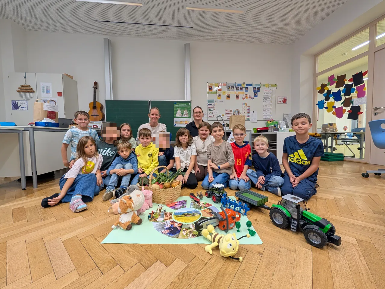 Eine Gruppe von Kindern und zwei Frauen sitzen auf dem Boden in einem Klassenzimmer. Es gibt Spielzeug, einen Korb mit Früchten und ein Poster an der Wand.