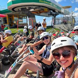 Eine Gruppe von Menschen mit Helmen und Sonnenbrillen fährt an einem sonnigen Tag auf Go-Karts vor einer Skilift.