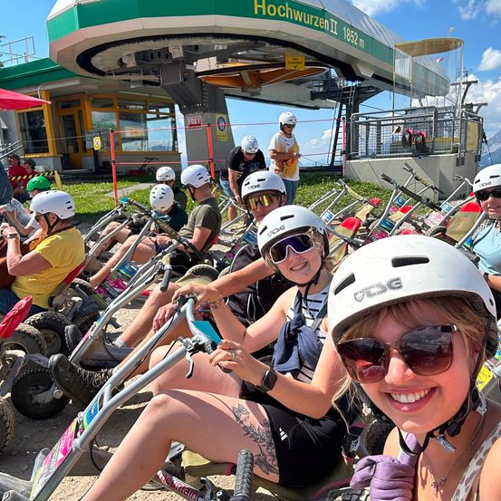 Eine Gruppe von Menschen mit Helmen und Sonnenbrillen fährt an einem sonnigen Tag auf Go-Karts vor einer Skilift.