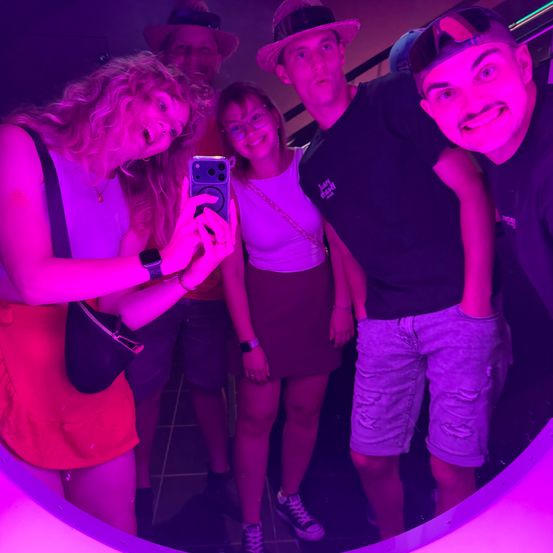 Eine Gruppe junger Erwachsener posiert für ein Foto in einem schwach beleuchteten Raum mit lila Beleuchtung. Eine Frau macht ein Selfie mit ihrem Handy, während andere lächeln.