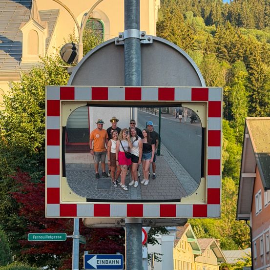 Eine Gruppe von Menschen steht auf einem gepflasterten Gehweg, reflektiert in einem Spiegel an einem Pfosten, mit einem Gebäude und Bäumen im Hintergrund.