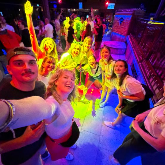 Eine Gruppe von Menschen in einem hell beleuchteten Club, einige mit gelber Gesichtsbemalung, tanzen und posieren für ein Foto.