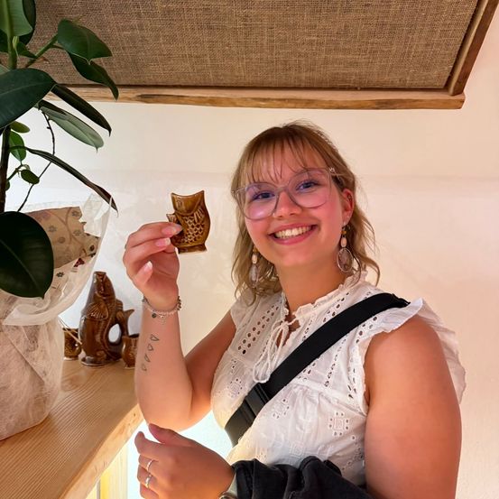 Eine Frau mit Brille hält eine hölzerne Fischfigur, lächelt und posiert für ein Foto in einem Raum mit einer Pflanze und einer Holzdecke.