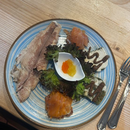 Ein Teller mit Fisch, Räucherlachs und einer kleinen Schale Kaviar auf einem Holztisch mit zwei Gabeln.