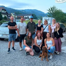 Eine Gruppe von Menschen und ein Hund posieren für ein Foto vor einer Bergkette, mit Häusern und Autos im Hintergrund.