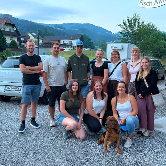Eine Gruppe von Menschen und ein Hund posieren für ein Foto vor einer Bergkette, mit Häusern und Autos im Hintergrund.