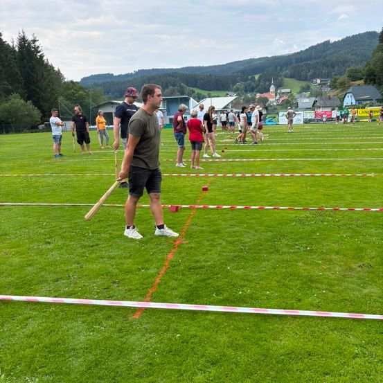 Eine Gruppe von Menschen spielt ein Spiel auf einem Rasenfeld mit roten und weißen Linien. Ein Mann hält einen Holzschläger.