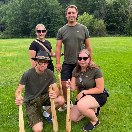 Vier Personen posieren mit Holzschlägern auf einem Rasenfeld für ein Foto. Sie lächeln und scheinen Spaß zu haben. Hinter ihnen befinden sich Bäume und ein Zaun.