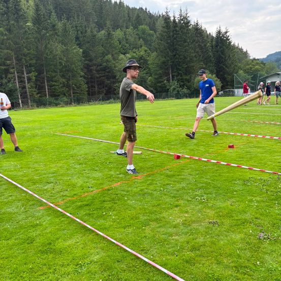 Eine Gruppe von Menschen spielt ein Spiel auf einem Rasenfeld mit einem großen Schläger, umgeben von Bäumen und Bergen.