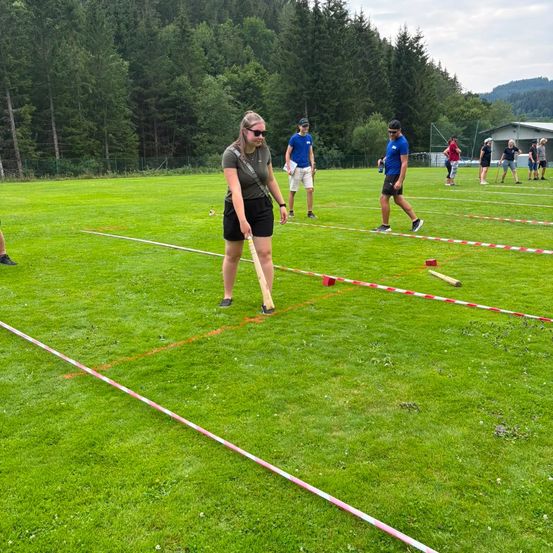 Eine Gruppe von Menschen spielt ein Spiel auf einem Rasenfeld, wobei eine Frau einen Stock hält und sich darauf vorbereitet, ihn zu werfen. Das Feld ist mit roten und weißen Linien markiert. Im Hintergrund befinden sich Bäume und ein Gebäude.