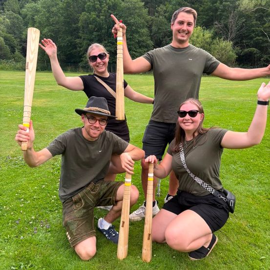 Vier Personen posieren für ein Foto auf einem Rasenfeld mit Holzschlägern, zwei knien und zwei stehen hinter ihnen. Sie alle lächeln und tragen Freizeitkleidung.