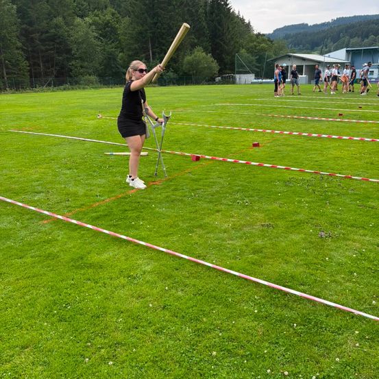 Eine Frau schwingt einen großen Holzschläger auf einem Rasenfeld, während eine Gruppe von Menschen im Hintergrund steht.