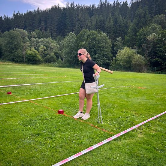 Eine Frau mit einer Krücke steht auf einem Rasenfeld, hält einen Schläger und rote und weiße Markierungen liegen auf dem Boden.