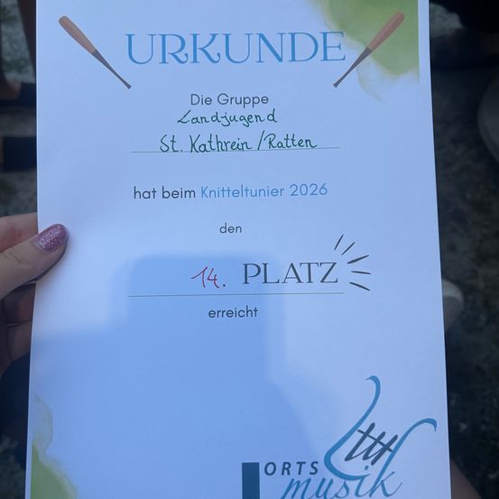 Ein von einer Hand gehaltenes Zertifikat zeigt ein blaues und grünes Design mit dem Text 'URKUNDE' und '14. PLATZ'. Es erwähnt 'Die Gruppe Landjugend St. Kathrein/Ratten' und 'hat beim Knitteltunier 2026 den'. Unten steht 'ORTS MUSIK'.
