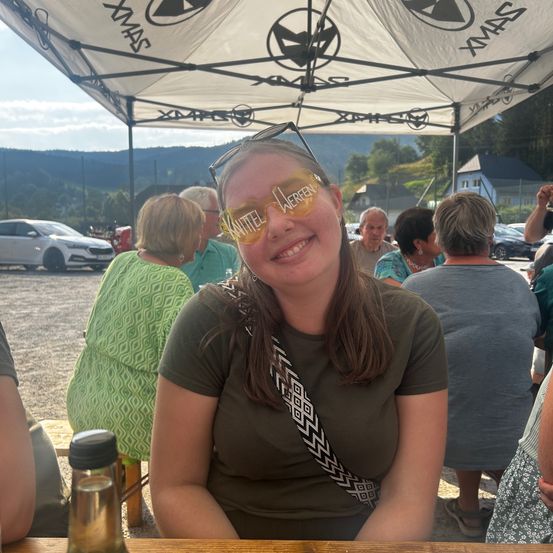 Eine Frau mit Sonnenbrille auf dem Kopf und einem Lächeln sitzt unter einem Zelt, umgeben von einer Gruppe von Menschen. Im Hintergrund sind geparkte Autos und eine Berglandschaft zu sehen.