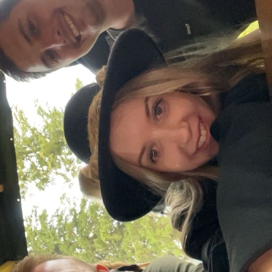 Eine Gruppe von Freunden, darunter eine Frau mit blonden Haaren und einem schwarzen Hut, lächelt und posiert für ein Foto in einer Außenumgebung mit Bäumen im Hintergrund.