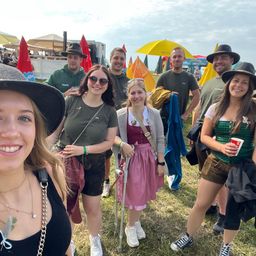 Eine Gruppe von Menschen, darunter Frauen in traditioneller bayerischer Kleidung, lächeln und posieren für ein Foto bei einer Veranstaltung im Freien. Einige tragen Hüte und halten Tassen. Im Hintergrund sind Zelte und Regenschirme zu sehen.